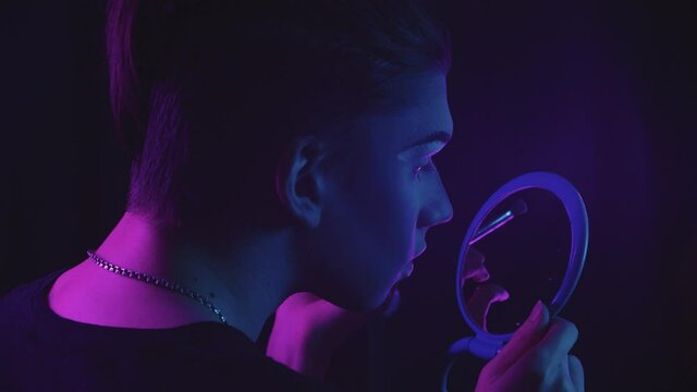 Drag artist - young man drawing new eyebrows and looking at himself in the mirror - blue and purple neon lighting