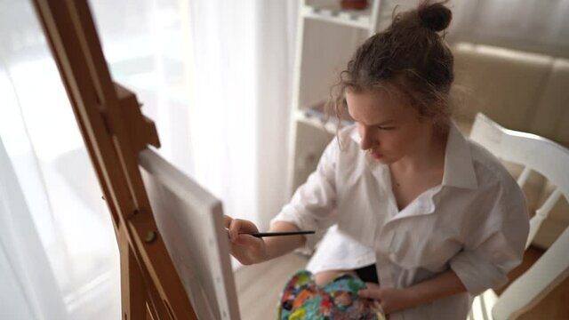 Young girl paints a portrait with oil paints at home. The schoolgirl sits on a chair near the easel and holds a palette and a brush in her hands. Children's leisure and creativity
