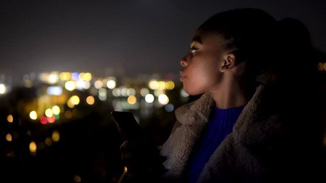 A young black woman looks around and at the smartphone in her hands in an urban area at night - closeup - screen light from smartphone illuminates her face - city lights in the blurry background