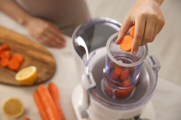 Young woman putting fresh slices of carrot into juicer at table in kitchen, above view