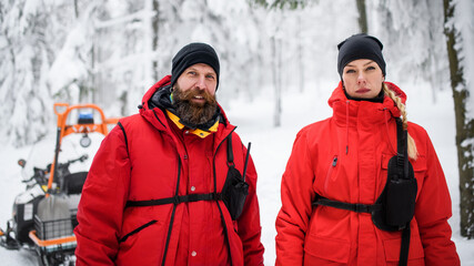 Woman and man paramedic from mountain rescue service outdoors in winter in forest, looking at camera.