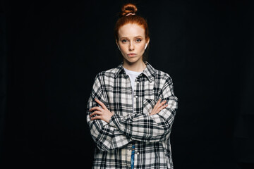 Serious red-haired young woman wearing wireless earphones with crossed hands looking at camera on isolated black background. Pretty redhead lady model emotionally showing facial expressions.