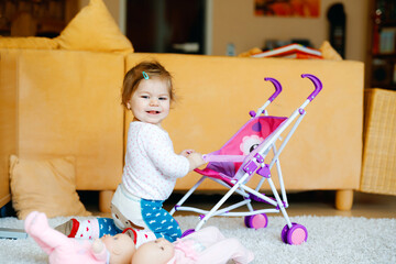 Cute adorable baby girl crawl and play with doll carriage. Beautiful toddler child with pushing...