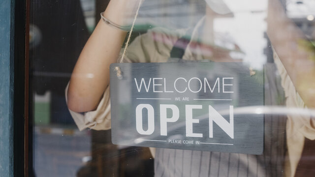 Young Asia Girl Wear Face Mask Turning A Sign From Closed To Open Sign On Door Looking Outside Waiting For Clients After Lockdown. Owner Small Business, Food And Drink, Business Reopen Again Concept.