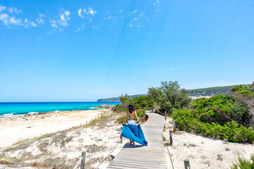Girl walking on a wooden walkway beside a beach with turquoise blue sea on a summer day.