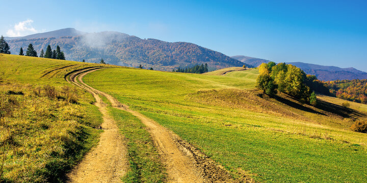mountainous countryside in autumn. rural road through grassy pastures on hills rolling in to the distance. forest in colorful foliage. bright sunny day with bright blue sky