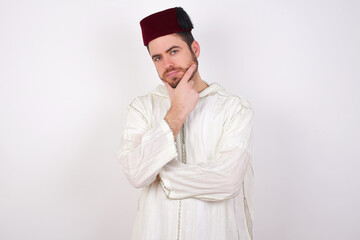 Thoughtful smiling young handsome Caucasian man wearing Arab djellaba and Fez hat over white wall keeps hand under chin, looks directly at camera, listens something with interest. Youth concept.