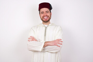 young handsome Caucasian man wearing Arab djellaba and Fez hat over white wall being happy smiling and crossed arms looking confident at the camera. Positive and confident person.