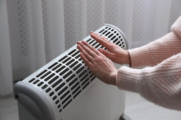 Woman warming hands near electric heater at home, closeup