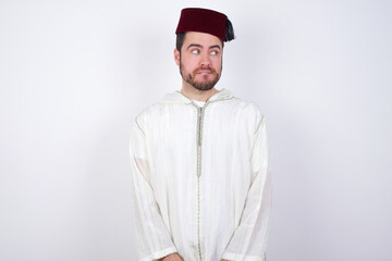 young handsome Caucasian man wearing Arab djellaba and Fez hat over white wall with thoughtful expression, looks away keeps hands down bitting his lip thinks about something pleasant.