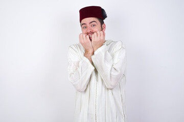 Anxiety - young handsome Caucasian man wearing Arab djellaba and Fez hat over white wall covering his mouth with hands scared from something or someone bitting nails.