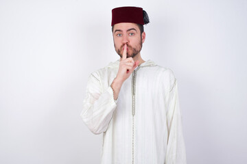 young handsome Caucasian man wearing Arab djellaba and Fez hat over white wall makes hush gesture, asks be quiet. Don't tell my secret or not speak too loud, please!