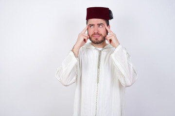 young handsome Caucasian man wearing Arab djellaba and Fez hat over white wall with thoughtful expression, looks away, keeps hand near face, thinks about something pleasant.