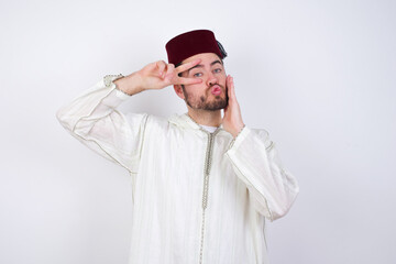 young handsome Caucasian man wearing Arab djellaba and Fez hat over white wall  making v-sign near eyes. Leisure lifestyle people person celebrate flirt coquettish concept.