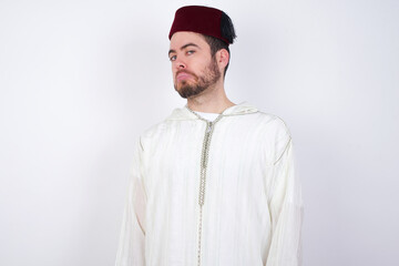 young handsome Caucasian man wearing Arab djellaba and Fez hat over white with snobbish expression curving lips and raising eyebrows, looking with doubtful and skeptical expression, suspect and doubt.