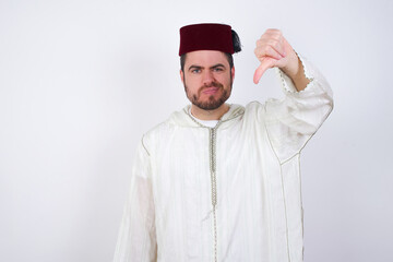 Obraz premium Discontent young handsome Caucasian man wearing Arab djellaba and Fez hat over white wall shows disapproval sign, keeps thumb down, expresses dislike, frowns face in discontent. Negative feelings.