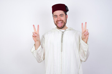 young handsome Caucasian man wearing Arab djellaba and Fez hat over white wall with optimistic smile, showing peace or victory gesture with both hands, looking friendly. V sign.