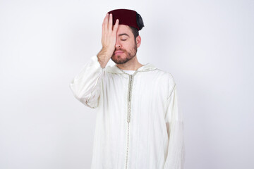 Frustrated young handsome Caucasian man wearing Arab djellaba and Fez hat over white wall holding hand on forehead being depressed regretting what he did having headache, looking stressful.