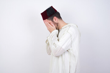 Sad young handsome Caucasian man wearing Arab djellaba and Fez hat over white wall covering face with hands and crying.