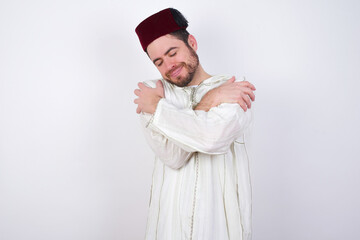 young handsome Caucasian man wearing Arab djellaba and Fez hat over white wall. Hugging oneself happy and positive, smiling confident. Self love and self care.