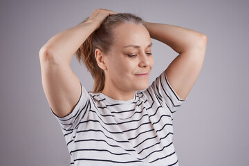 Mature woman in a striped T-shirt makes a ponytail hairstyle, looks down. Studio shot against gray background.