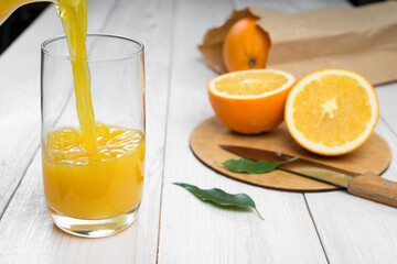 Orange juice is poured into a glass on a wooden table, fresh cut oranges in the background