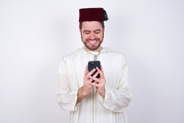 Smiling young handsome Caucasian man wearing Arab djellaba and Fez hat using cell phone, messaging, being happy to text with friends, looking at smartphone. Modern technologies and communication.