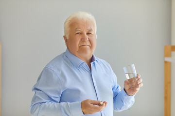 Positive senior man in blue shirt holding medical pill and pure water in glass in hands