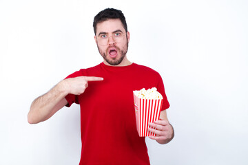 young handsome man in red T-shirt against white background eating popcorn being in stupor shocked,...