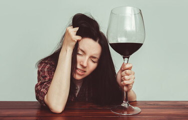 A young caucasian girl in a plaid shirt with tousled hair sits at a wooden table and holds a large glass of red wine.Concept of alcohol abuse, headache, alcoholism, hangover, loneliness and depression