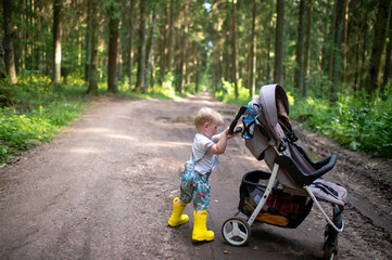 Fototapeta na wymiar A little funny boy in yellow boots posing with the baby carriage in the forest