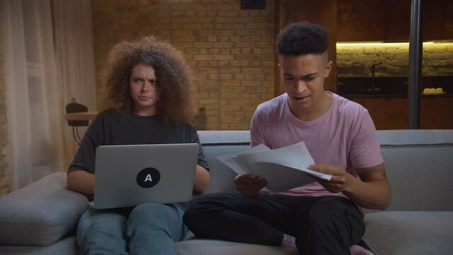 Young African American man giving high five to girlfriend after passing online exam. Young mixed race couple cheating while online interview for new job sitting on sofa.