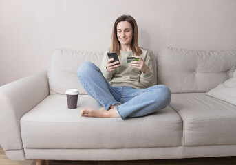 young woman sitting on sofa enjoying shopping in mobile application, entering information from banking credit card, confirming purchase of delivery , online order.