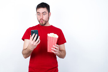 Young handsome man in red T-shirt against white background eating popcorn looks with bugged eyes, holds modern smart phone, receives unexpected message from friend, reads reminder.