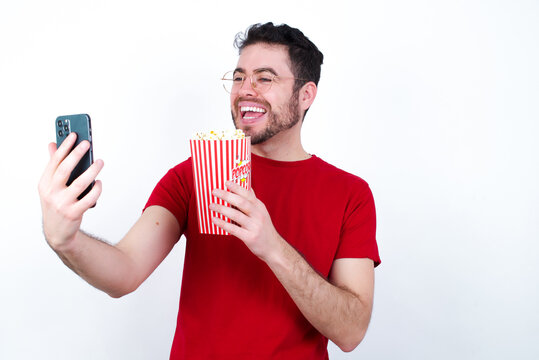 Portrait of happy friendly Young handsome man in red T-shirt against white background eating popcorn taking selfie and waving hand, communicating on video call, online chatting.