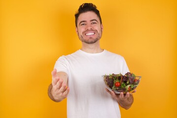 young handsome Caucasian man holding a salad bowl against yellow wall smiling friendly offering something with open hand or handshake as greeting and welcoming. Successful business.
