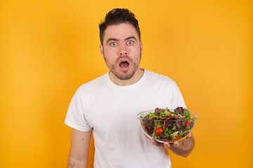 young handsome Caucasian man holding a salad bowl against yellow wall having stunned and shocked look, with mouth open and jaw dropped exclaiming: Wow, I can't believe this. Surprise and shock