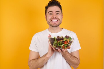 young handsome Caucasian man holding a salad bowl against yellow wall keeps palms together, has pleased expression. Glad attractive male makes request, pleads for mercy. Hopeful young adult.
