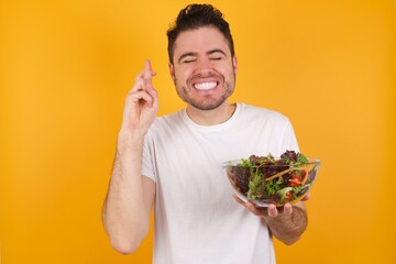 Joyful young handsome Caucasian man holding a salad bowl against yellow wall clenches teeth, raises fingers crossed, makes desirable wish, waits for good news, I have to win.