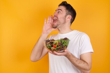 young handsome Caucasian man holding a salad bowl against yellow wall profile view, looking happy and excited, shouting and calling to copy space.