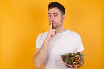 young handsome Caucasian man holding a salad bowl against yellow wall makes hush gesture, asks be...