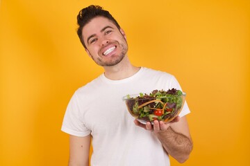 young handsome Caucasian man holding a salad bowl against yellow wall with broad smile, shows white teeth, feeling confident rejoices having day off.