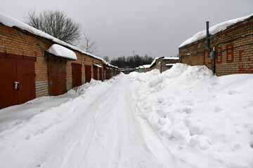 A cloudy winter day. Snowfall. Rows of one-story brick garages with closed metal painted gates. Drifts of snow near the walls along the road. Snow on the roofs.