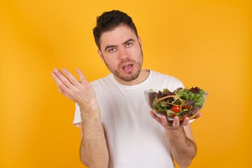 Studio shot of frustrated young handsome Caucasian man holding a salad bowl against yellow wall gesturing with raised palm, frowning, being displeased and confused with dumb question.