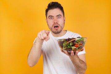Shocked young handsome Caucasian man holding a salad bowl against yellow wall points front with index finger at camera and. Surprise and advertisement concept.