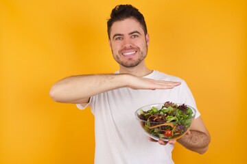 young handsome Caucasian man holding a salad bowl against yellow wall gesturing with hands showing big and large size sign, measure symbol.