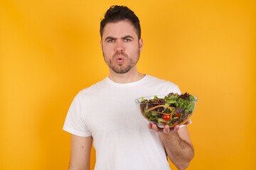 young handsome Caucasian man holding a salad bowl against yellow wall expressing disgust, unwillingness, disregard having tensive look frowning face, looking indignant with something.