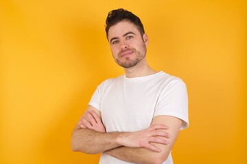 Waist up shot of  self confident young handsome Caucasian man against yellow wall has broad smile, crosses arms, happy to meet with colleagues.