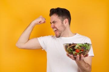 Yes I am winner. Portrait of charming delighted and excited young handsome Caucasian man holding a salad bowl raising up fist in triumph and victory smiling achieving success grinning from delight.