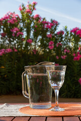 Jug and glass with water on a table with tablecloth on the outside porch of a house with a garden and flowers in the background